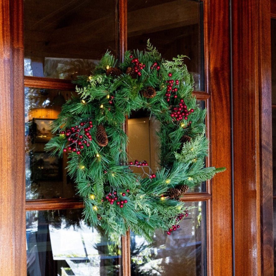 A pre-lit Plymouth wreath glowing warm white on a mahogany-framed glass door at dusk