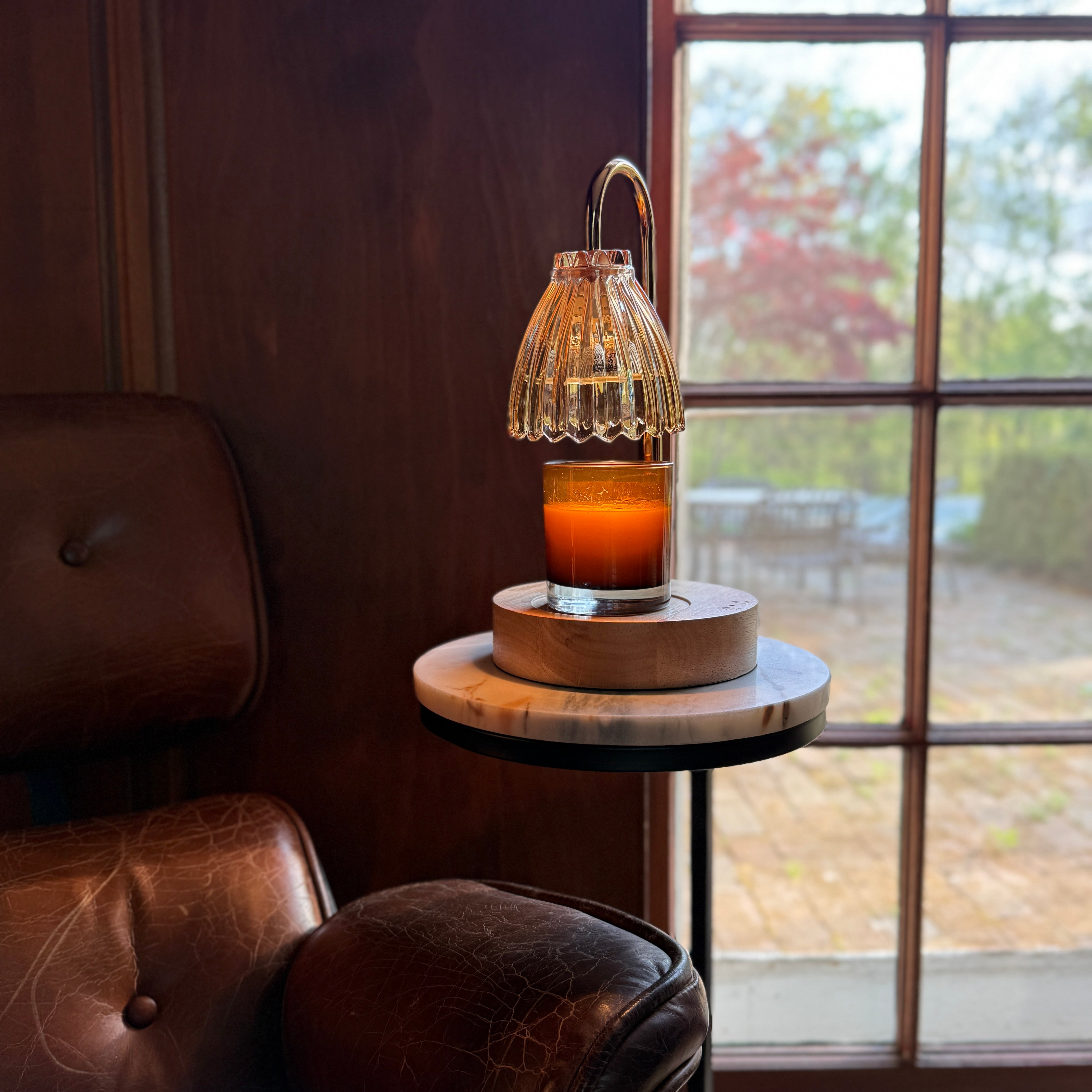 Amber large jar glass candle resting on a cordless candle warmer that has a clear, tulip shade glass bulb, light wood base, resting on a small side table next to an accent chair in living room.