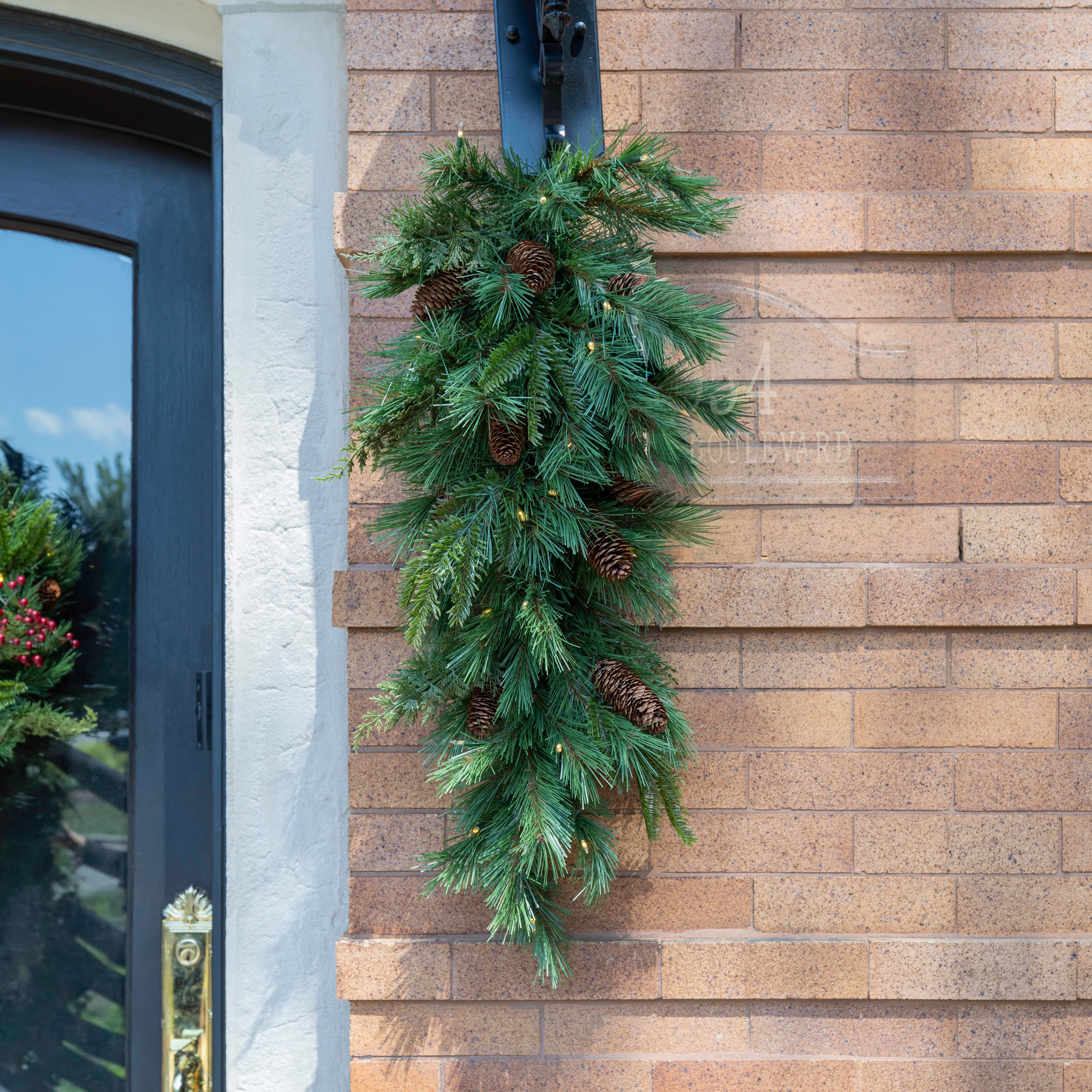 Woodsy, evergreen teardrop arrangement with cedar and pine tips and real pinecone clusters. Pre-lit with warm white lights. Hanging off a lantern against a red brick home.