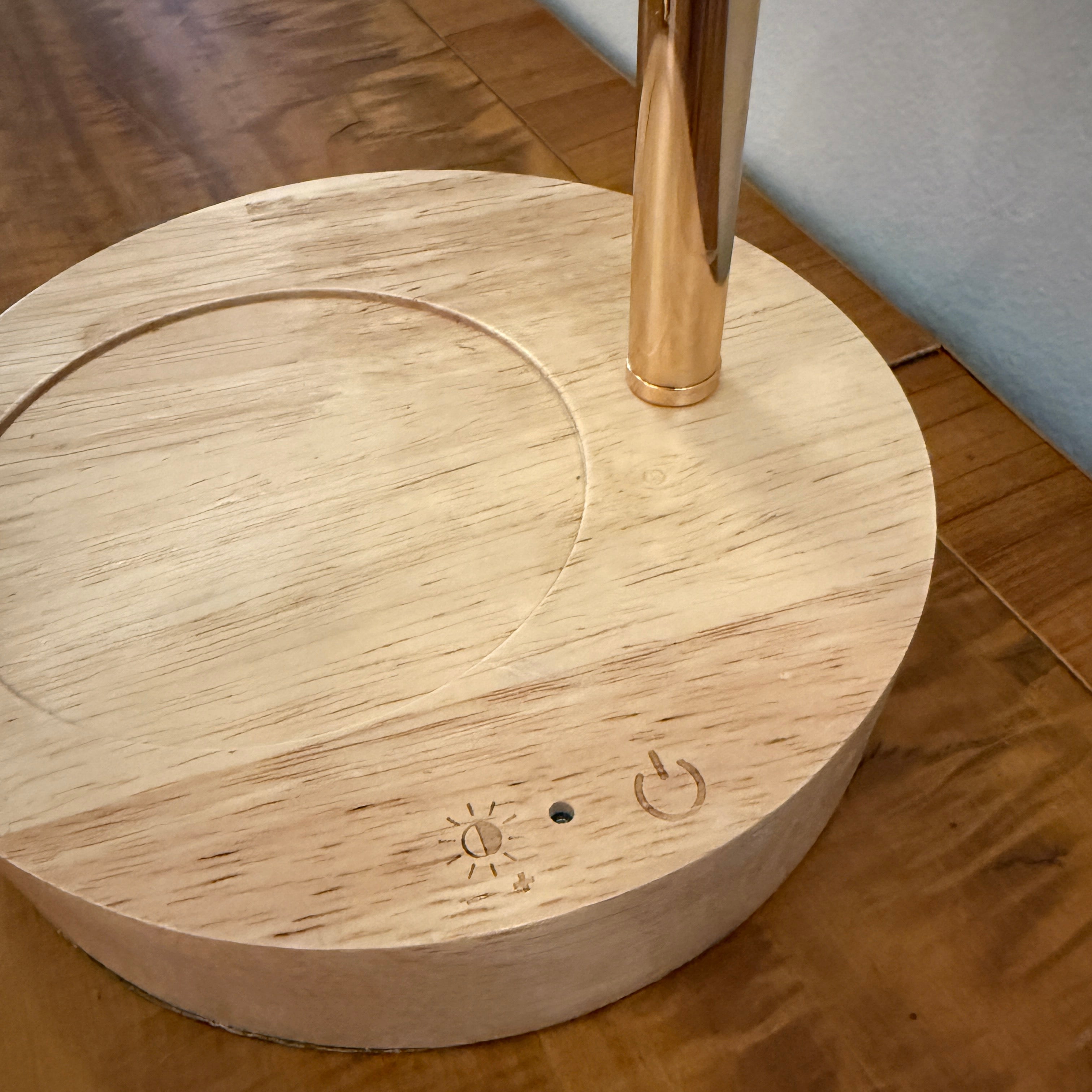 A close-up of a round light-wood base with engraved controls and a vertical metallic stem, placed on a wooden floor.