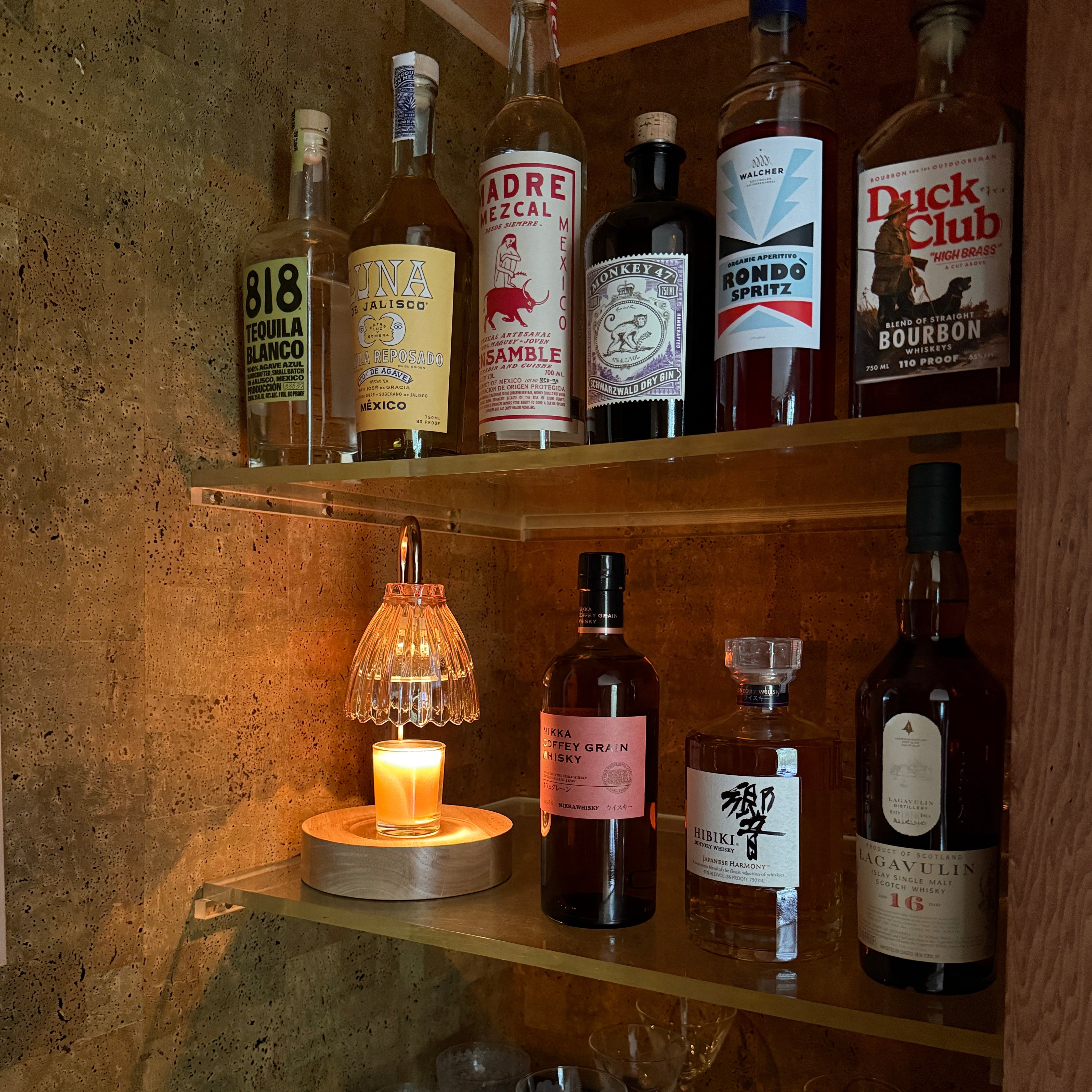 A warm, dimly lit home bar corner with wooden shelves holding an assortment of liquor bottles, a lit candle in a glass holder, and several wine glasses on a dark counter against a textured wall.