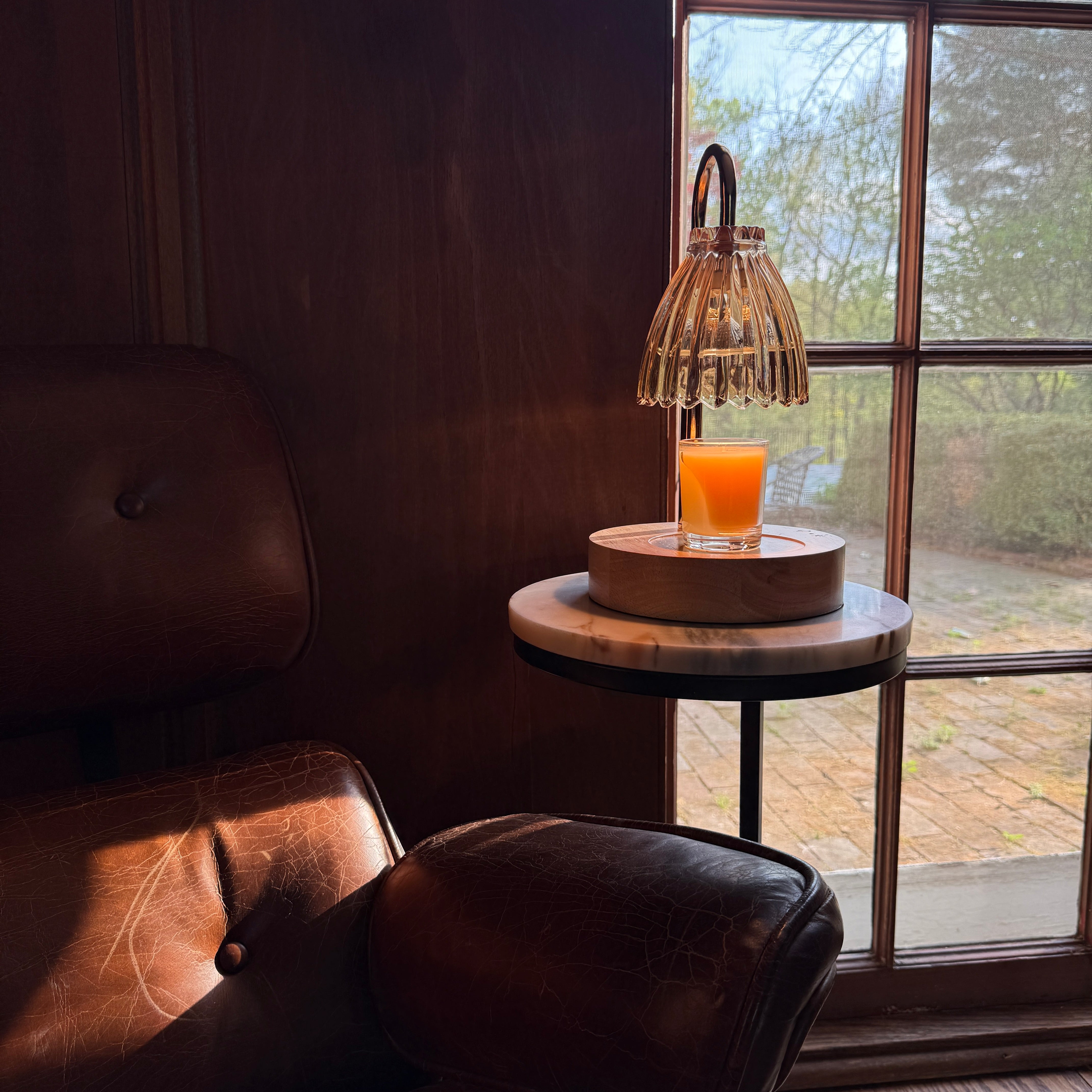 A modern glass table lamp with a ribbed bell shade and curved handle, illuminated and placed on a small round marble side table next to a leather armchair in a warm, sunlit room.