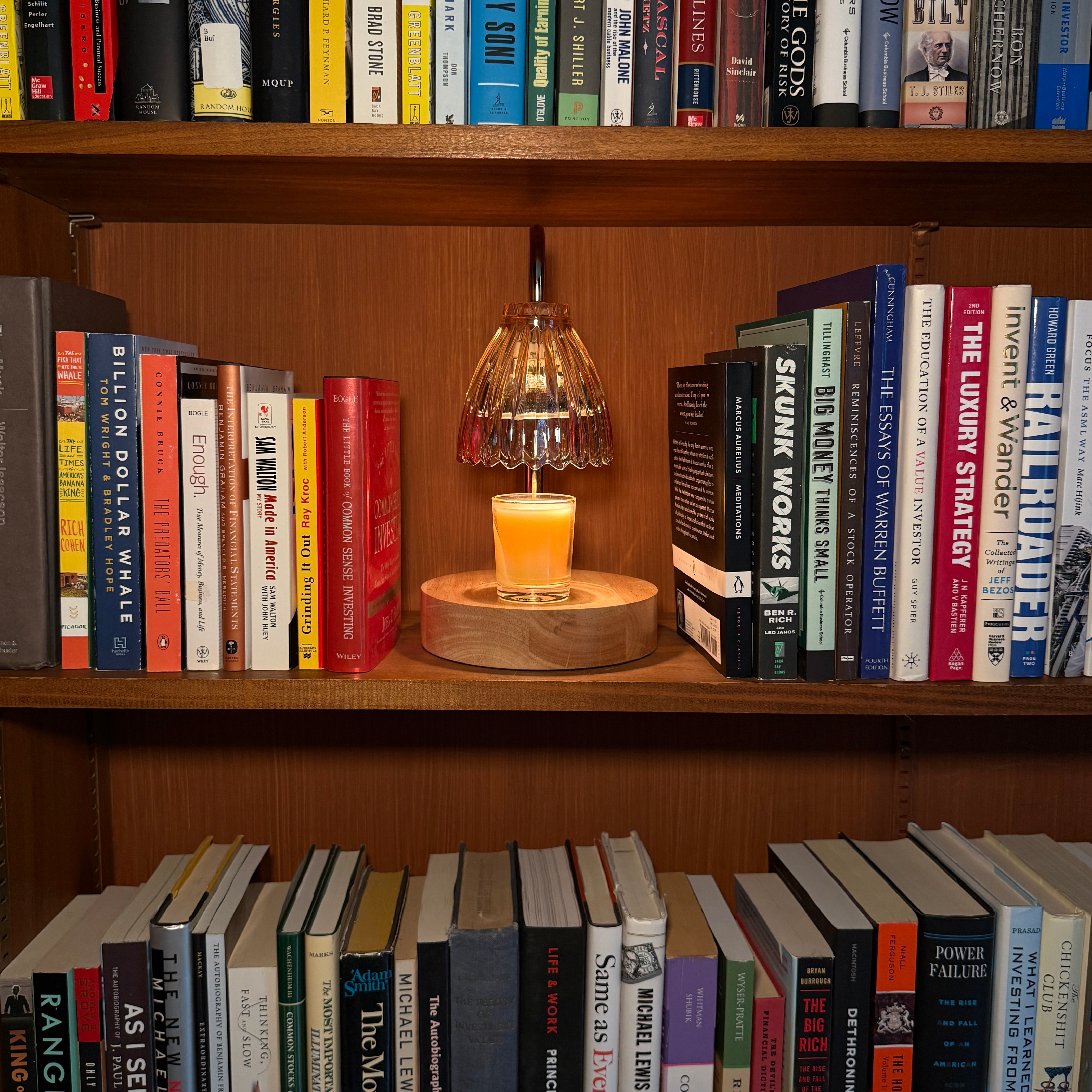 A small candle on a cordless candle warmer with a glass shade with a wooden base sits on a wooden bookshelf between rows of books.
