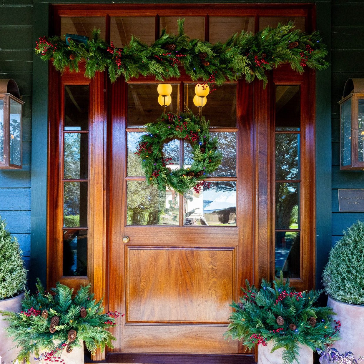 Christmas entryway with a lit wreath on the door, a matching garland swag above, and berry-filled urns in stone pots flanking the steps
