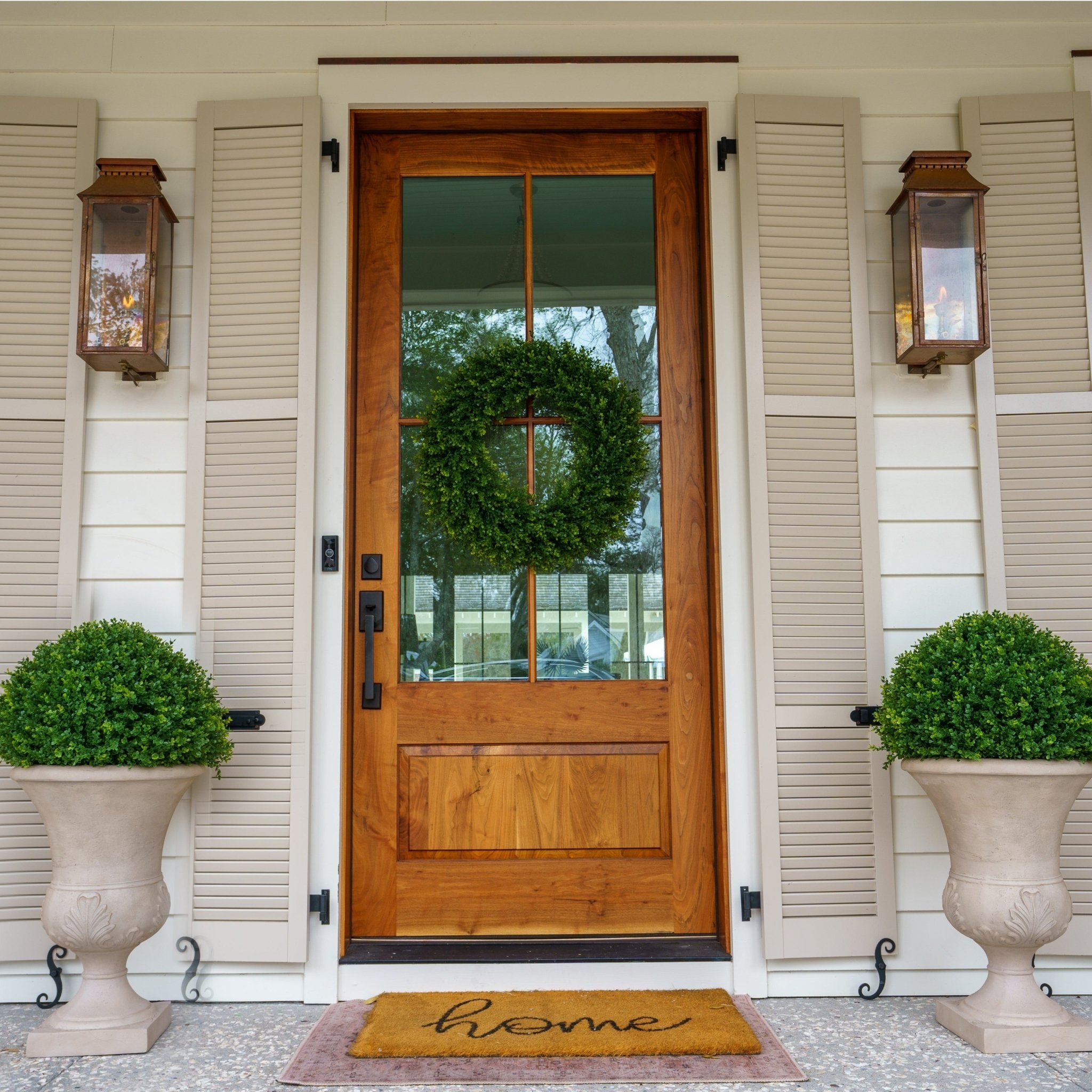 Matched artificial boxwood spheres in stone urns flanking a glass-paneled wood front door with a boxwood wreath