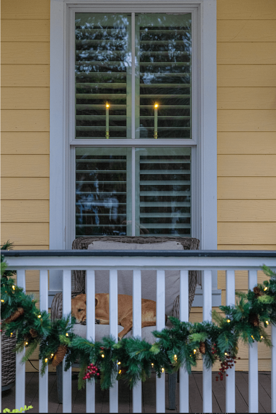 Front porch at dusk with battery window candles glowing in the window and pre-lit garland on the railing, no cords visible